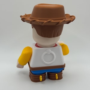 Op de afbeelding: Een 3D-geprint figuur van een cartoon cowboy-personage, van achteren bekeken. Het figuur draagt een bruine cowboyhoed, een wit shirt met een ringdetail, een blauwe broek, bruine laarzen en gele mouwen. Het figuur staat op een witte achtergrond.