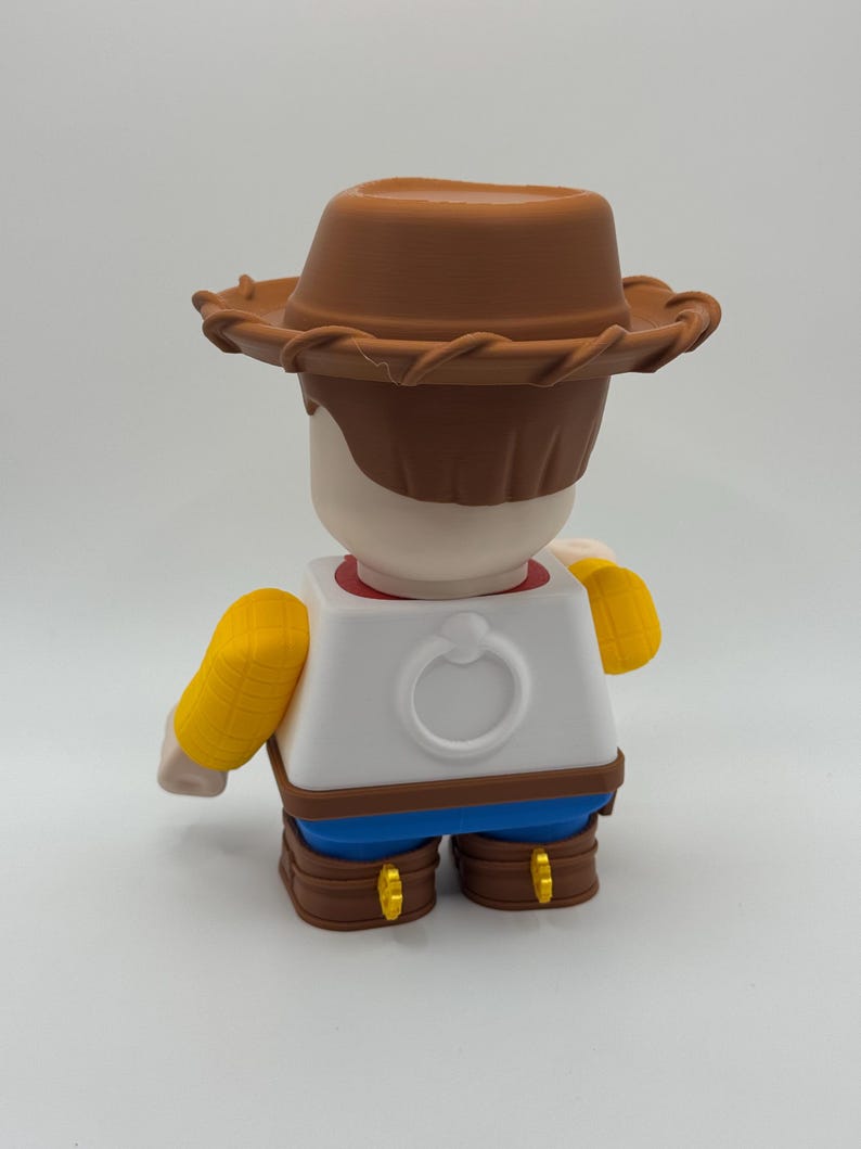 Op de afbeelding: Een 3D-geprint figuur van een cartoon cowboy-personage, van achteren bekeken. Het figuur draagt een bruine cowboyhoed, een wit shirt met een ringdetail, een blauwe broek, bruine laarzen en gele mouwen. Het figuur staat op een witte achtergrond.