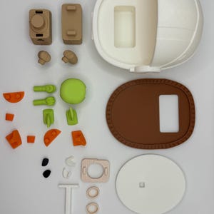 Puede incluir: Un conjunto de juguetes desmontados de varios colores. El conjunto incluye una estructura esf&eacute;rica blanca, una base ovalada marr&oacute;n y piezas m&aacute;s peque&ntilde;as en tonos marr&oacute;n, verde, naranja y blanco. Las piezas parecen estar impresas en 3D, probablemente para un juguete infantil.