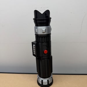 Puede incluir: Un accesorio de sable de luz negro y plateado con un bot&oacute;n rojo y un dise&ntilde;o superior &uacute;nico. El accesorio es cil&iacute;ndrico con crestas detalladas y una secci&oacute;n media plateada. Es una r&eacute;plica de un arma de ciencia ficci&oacute;n popular.