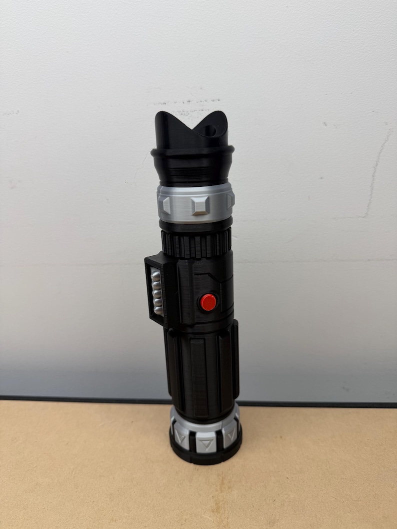 Puede incluir: Un accesorio de sable de luz negro y plateado con un bot&oacute;n rojo y un dise&ntilde;o superior &uacute;nico. El accesorio es cil&iacute;ndrico con crestas detalladas y una secci&oacute;n media plateada. Es una r&eacute;plica de un arma de ciencia ficci&oacute;n popular.