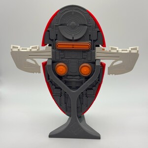 Puede incluir: Modelo detallado de una nave espacial, con un cuerpo rojo y gris y detalles en naranja y blanco. El modelo est&aacute; montado sobre un soporte gris, mostrando su intrincado dise&ntilde;o y estructura sim&eacute;trica. Las alas est&aacute;n extendidas.