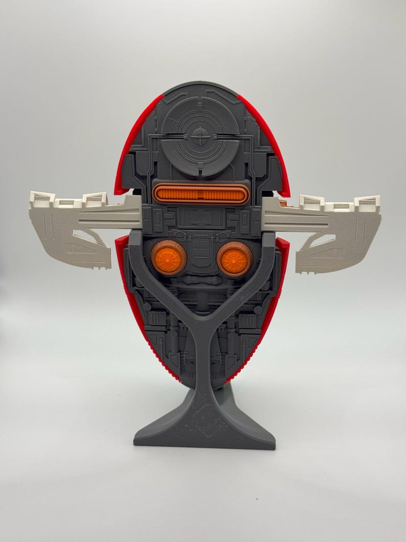 Puede incluir: Modelo detallado de una nave espacial, con un cuerpo rojo y gris y detalles en naranja y blanco. El modelo est&aacute; montado sobre un soporte gris, mostrando su intrincado dise&ntilde;o y estructura sim&eacute;trica. Las alas est&aacute;n extendidas.
