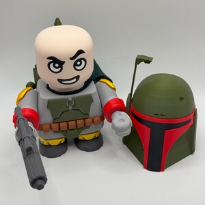 Puede incluir: Una figura impresa en 3D de un personaje con cabeza calva, capa verde y atuendo gris y rojo, sosteniendo un bl&aacute;ster gris. Junto a &eacute;l, un casco verde con visera roja. La figura tiene una expresi&oacute;n severa.