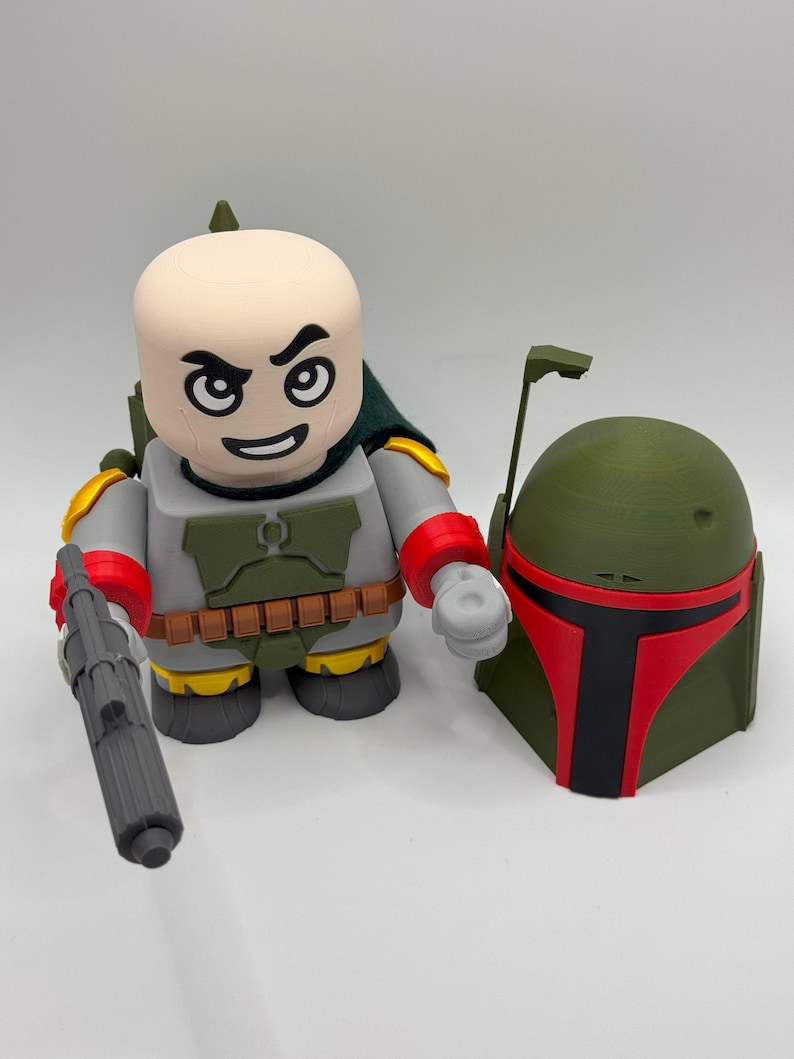 Puede incluir: Una figura impresa en 3D de un personaje con cabeza calva, capa verde y atuendo gris y rojo, sosteniendo un bl&aacute;ster gris. Junto a &eacute;l, un casco verde con visera roja. La figura tiene una expresi&oacute;n severa.