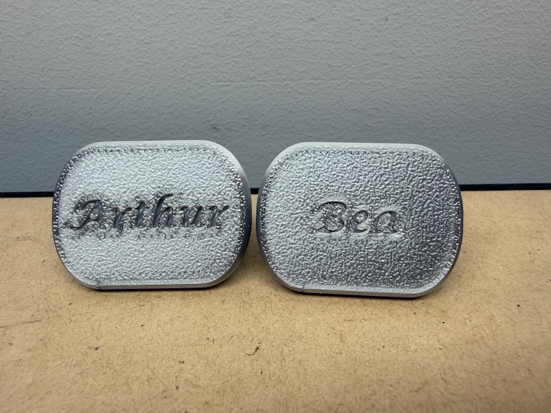 May include: Two oval-shaped silver-colored objects with a textured surface. The left object features the name Arthur embossed in a cursive script, while the right object features the name Bea embossed in a similar cursive script style.