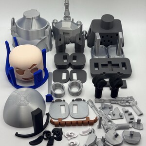 Peut inclure: Diverses pi&egrave;ces imprim&eacute;es en 3D, de couleurs grises, noires et beiges. L'ensemble comprend des &eacute;l&eacute;ments pour un casque, des armes et d'autres composants m&eacute;caniques. Les pi&egrave;ces sont dispos&eacute;es sur une surface blanche. Mesures non applicables.
