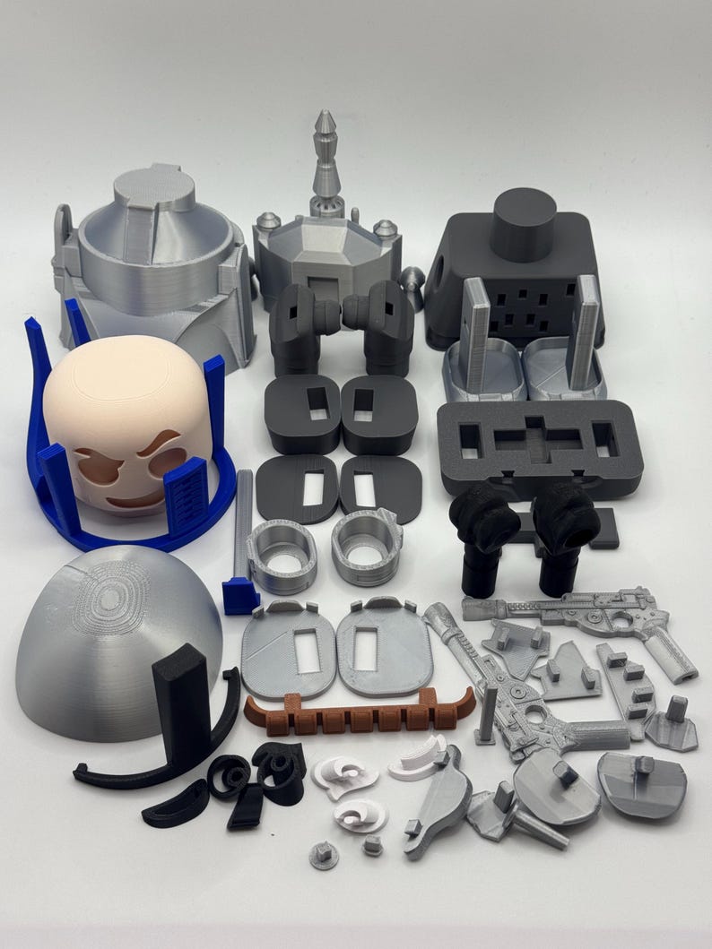Peut inclure: Diverses pi&egrave;ces imprim&eacute;es en 3D, de couleurs grises, noires et beiges. L'ensemble comprend des &eacute;l&eacute;ments pour un casque, des armes et d'autres composants m&eacute;caniques. Les pi&egrave;ces sont dispos&eacute;es sur une surface blanche. Mesures non applicables.