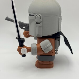 May include: A grey and brown action figure of a Mandalorian with a black lightsaber and a blaster pistol. The figure is wearing a helmet and a cape.