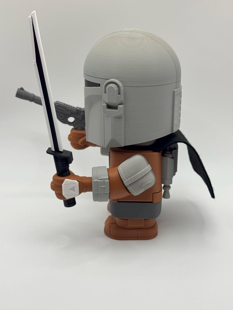 May include: A grey and brown action figure of a Mandalorian with a black lightsaber and a blaster pistol. The figure is wearing a helmet and a cape.