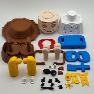 Op de afbeelding: Een verzameling kleurrijke 3D-geprinte speelgoedonderdelen, waaronder een bruine cowboyhoed, een beige gezicht met een lachende uitdrukking en diverse andere componenten in blauw, geel en wit. De onderdelen zijn gerangschikt op een witte ondergrond.