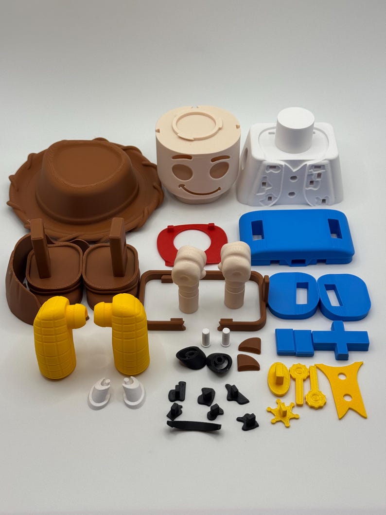 Op de afbeelding: Een verzameling kleurrijke 3D-geprinte speelgoedonderdelen, waaronder een bruine cowboyhoed, een beige gezicht met een lachende uitdrukking en diverse andere componenten in blauw, geel en wit. De onderdelen zijn gerangschikt op een witte ondergrond.