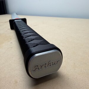 May include: A sword handle featuring a black textured grip with a cross-hatched pattern and a rectangular silver-colored pommel. The pommel is engraved with the name Arthur in a cursive script. A black guard separates the handle from the blade.