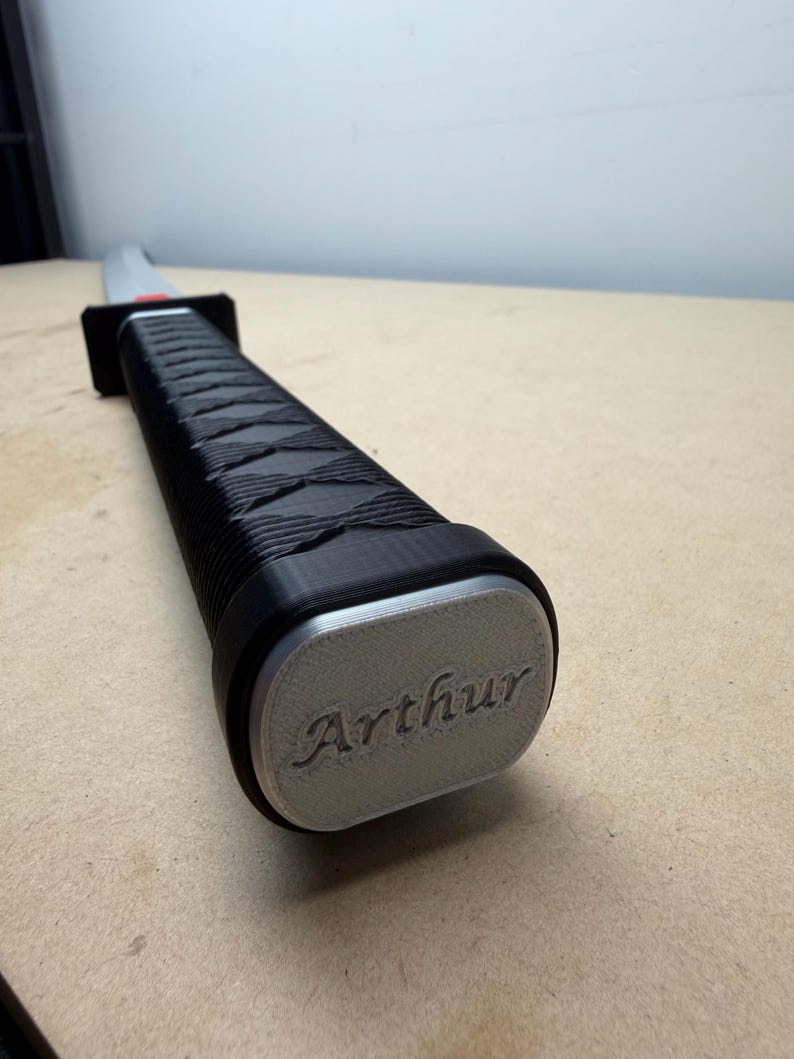 May include: A sword handle featuring a black textured grip with a cross-hatched pattern and a rectangular silver-colored pommel. The pommel is engraved with the name Arthur in a cursive script. A black guard separates the handle from the blade.