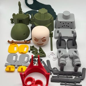 Puede incluir: Un surtido de piezas de juguetes impresas en 3D en varios colores, incluyendo verde oliva, gris, amarillo, rojo y beige. La colecci&oacute;n presenta componentes que se asemejan a un tanque, armas y otros elementos de construcci&oacute;n. Las piezas est&aacute;n dispuestas sobre una superficie blanca.