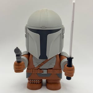 May include: A 3D printed action figure of a Mandalorian character from the Star Wars universe. The figure is wearing a grey helmet and brown armour, and is holding a blaster pistol in one hand and a lightsaber in the other.