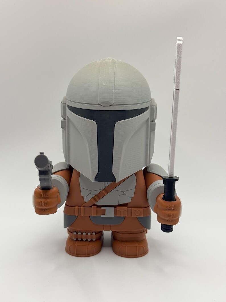 May include: A 3D printed action figure of a Mandalorian character from the Star Wars universe. The figure is wearing a grey helmet and brown armour, and is holding a blaster pistol in one hand and a lightsaber in the other.