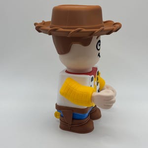 Op de afbeelding: Een grote, gestileerde figuur van een cowboy-personage, met een bruine hoed, bruin haar en een wit overhemd met een rode halsdoek. De figuur heeft gele mouwen, een blauwe broek en bruine laarzen. De figuur staat op een wit oppervlak.