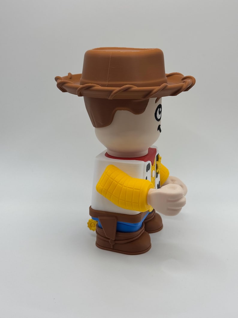 Op de afbeelding: Een grote, gestileerde figuur van een cowboy-personage, met een bruine hoed, bruin haar en een wit overhemd met een rode halsdoek. De figuur heeft gele mouwen, een blauwe broek en bruine laarzen. De figuur staat op een wit oppervlak.