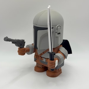 May include: A grey and brown action figure of a Mandalorian character from the Star Wars franchise. The figure is holding a blaster pistol in one hand and a lightsaber in the other. The figure is wearing a helmet and armour.