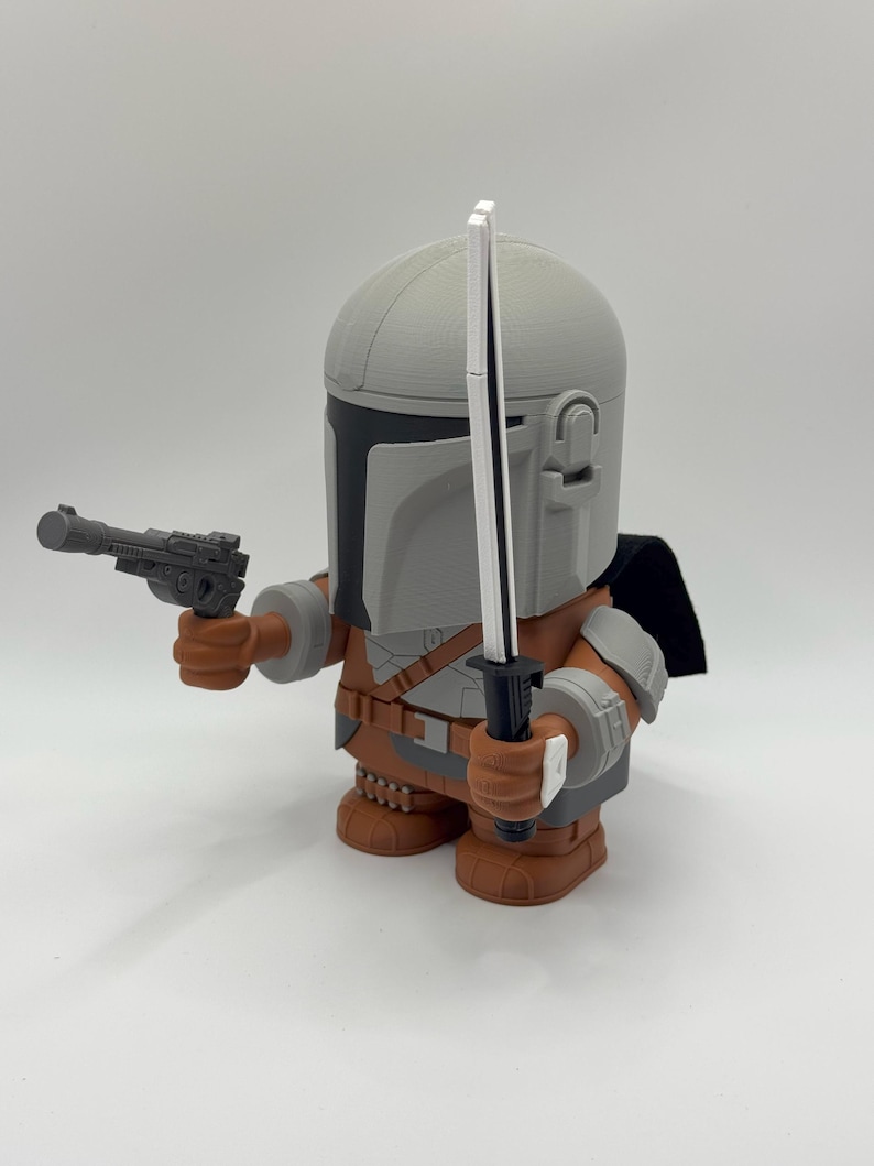 May include: A grey and brown action figure of a Mandalorian character from the Star Wars franchise. The figure is holding a blaster pistol in one hand and a lightsaber in the other. The figure is wearing a helmet and armour.