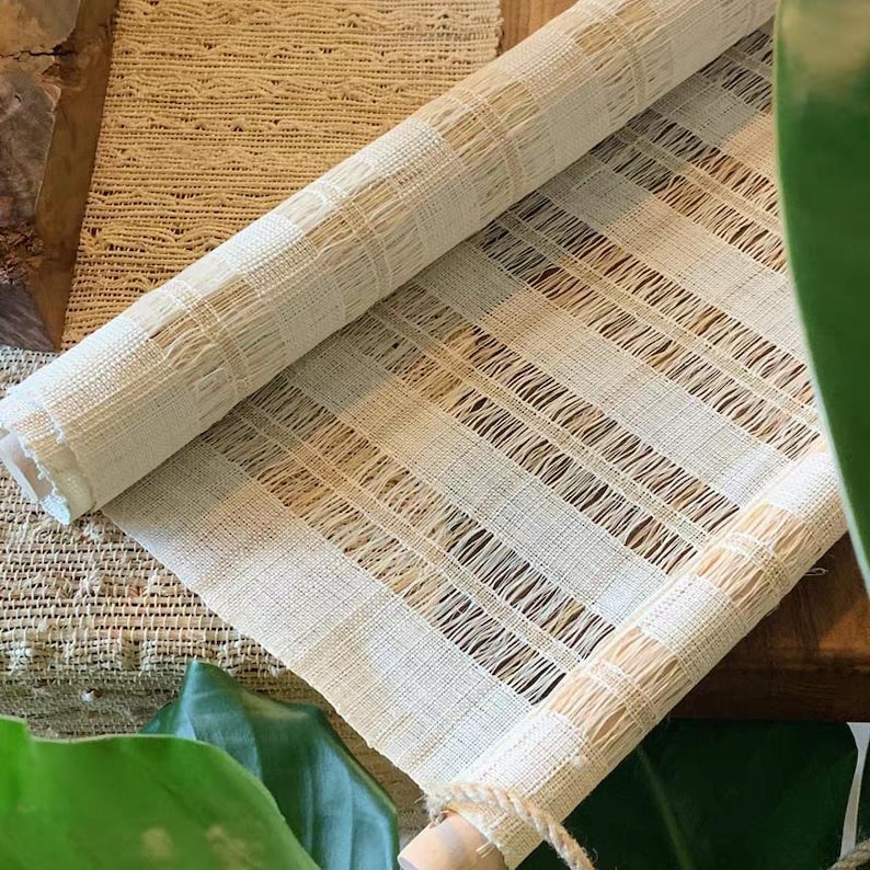 Puede incluir: Un camino de mesa de tela tejida de color crema con un patr&oacute;n de rayas texturizado y semitransparente, con secciones alternas s&oacute;lidas y de tejido abierto. El camino est&aacute; enrollado en ambos extremos, mostrando una estructura textil ligera.