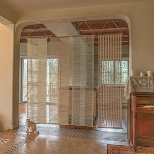 Può includere: Divisori per ambienti in tessuto beige appesi al soffitto, creando un'estetica naturale e ariosa. I divisori sono realizzati in un materiale chiaro e strutturato, con motivi diversi. Un gatto è seduto sul pavimento davanti ai divisori. La stanza ha un soffitto in legno e un pianoforte.