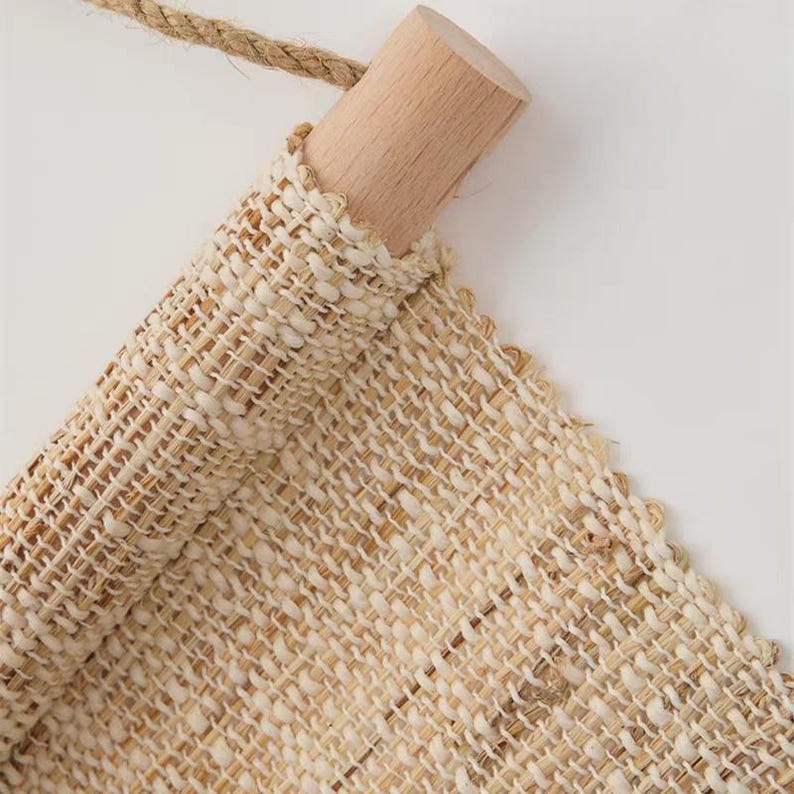 Puede incluir: Un tapiz de tela tejida en color beige claro suspendido de un cilindro de madera de color claro. El tejido presenta una textura gruesa con fibras naturales visibles y est&aacute; sujeto a una cuerda de yute delgada y trenzada para colgarlo.