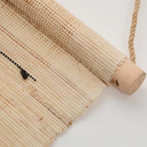 Puede incluir: Un tapiz de tela beige tejida con un cilindro de madera clara en la parte superior. Una cuerda de yute gruesa de tono natural est&aacute; sujeta al cilindro para colgarlo. La tela muestra una textura de tejido abierto con l&iacute;neas negras finas.