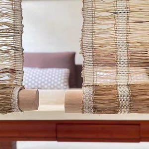 Puede incluir: Dos persianas verticales de ca&ntilde;a tejida con un patr&oacute;n de rayas en beige y marr&oacute;n natural. Cada persiana est&aacute; enrollada en la parte inferior alrededor de un cilindro de madera lisa de color claro, creando un cubrimiento de ventana texturizado y semitransparente.