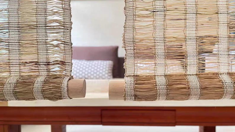Puede incluir: Dos persianas verticales de ca&ntilde;a tejida con un patr&oacute;n de rayas en beige y marr&oacute;n natural. Cada persiana est&aacute; enrollada en la parte inferior alrededor de un cilindro de madera lisa de color claro, creando un cubrimiento de ventana texturizado y semitransparente.