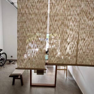 Puede incluir: Dos cortinas de ventana rectangulares hechas de fibras naturales tejidas con un patr&oacute;n vertical abstracto en color marr&oacute;n claro y crema. Las cortinas cuelgan una al lado de la otra, filtrando parcialmente la luz y permitiendo ver la habitaci&oacute;n.