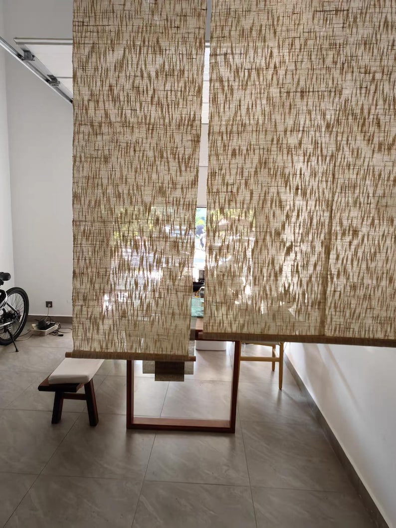 Puede incluir: Dos cortinas de ventana rectangulares hechas de fibras naturales tejidas con un patr&oacute;n vertical abstracto en color marr&oacute;n claro y crema. Las cortinas cuelgan una al lado de la otra, filtrando parcialmente la luz y permitiendo ver la habitaci&oacute;n.