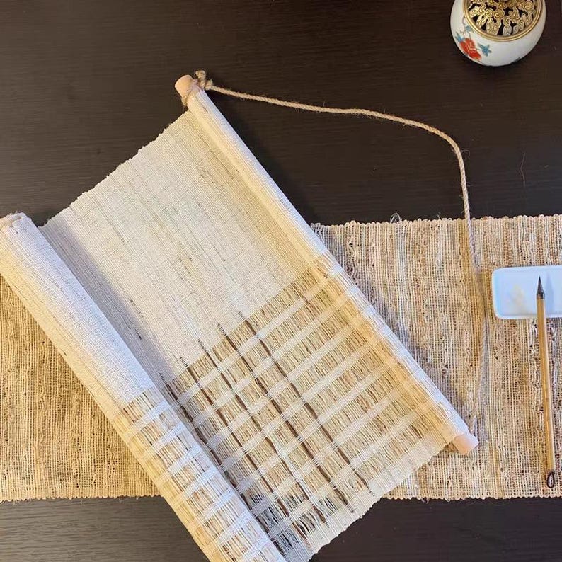 Puede incluir: Un tapiz de pared tejido en color beige con un panel de tela claro que presenta una textura de rejilla en la parte inferior, suspendido por una cuerda de yute fina. Un pincel de madera con punta oscura descansa sobre una bandeja blanca.