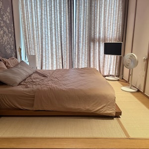 Pu&ograve; includere: Camera da letto con un letto basso in legno, coperto da un piumino e cuscini rosa cipria. Un tappetino tatami beige chiaro copre il pavimento. Una TV a schermo piatto e un ventilatore si trovano vicino alla finestra con tende trasparenti. Carta da parati floreale.