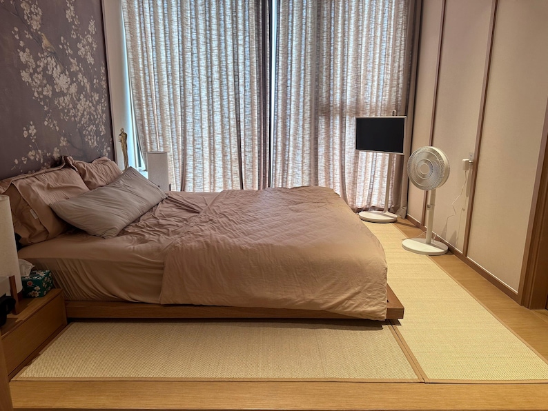 Pu&ograve; includere: Camera da letto con un letto basso in legno, coperto da un piumino e cuscini rosa cipria. Un tappetino tatami beige chiaro copre il pavimento. Una TV a schermo piatto e un ventilatore si trovano vicino alla finestra con tende trasparenti. Carta da parati floreale.