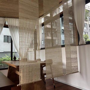 Puede incluir: Tres separadores de ambientes colgantes verticales hechos de un material de fibra natural tejida en color beige claro. Cada panel presenta un patr&oacute;n de rejilla semitransparente y texturizado, con una varilla de madera en la base.