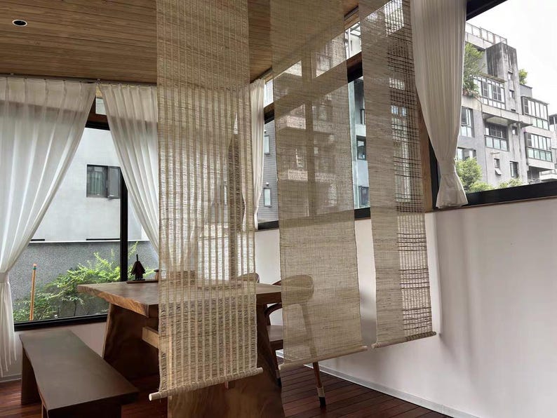 Puede incluir: Tres separadores de ambientes colgantes verticales hechos de un material de fibra natural tejida en color beige claro. Cada panel presenta un patr&oacute;n de rejilla semitransparente y texturizado, con una varilla de madera en la base.