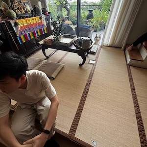 Pu&ograve; includere: Una stanza con stuoie tatami, un tavolino basso in legno e una grande finestra con vista su un giardino. Una persona &egrave; seduta sul pavimento. Un cane &egrave; visibile vicino alla finestra. Oggetti decorativi sono sul tavolo e sullo sfondo.