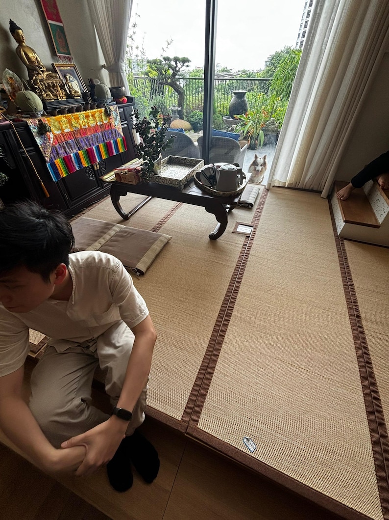 Pu&ograve; includere: Una stanza con stuoie tatami, un tavolino basso in legno e una grande finestra con vista su un giardino. Una persona &egrave; seduta sul pavimento. Un cane &egrave; visibile vicino alla finestra. Oggetti decorativi sono sul tavolo e sullo sfondo.