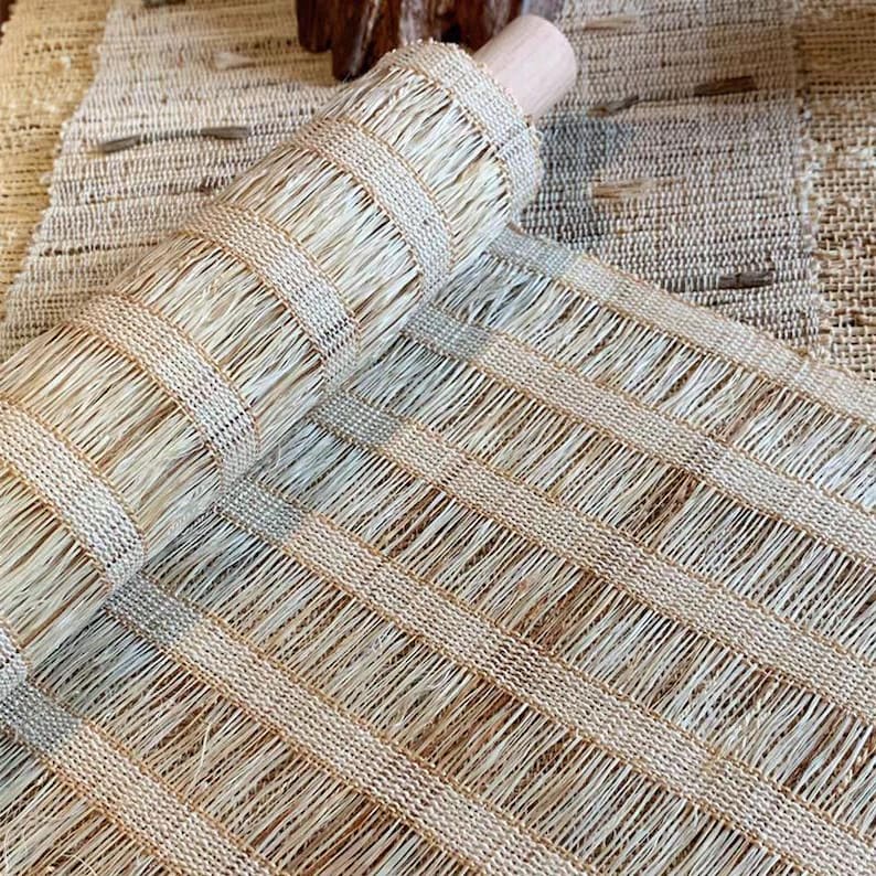 Puede incluir: Un rollo de esterilla de fibra natural tejida con un patr&oacute;n de cuadr&iacute;cula de hebras verticales y horizontales en color beige claro y tostado. El material tiene una apariencia fibrosa y texturizada con una estructura de tejido visible.