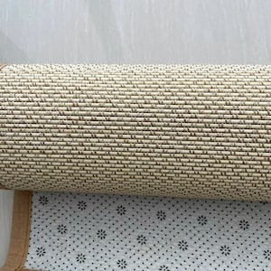 Pu&ograve; includere: Tappeto intrecciato beige e marrone arrotolato con un bordo marrone. Il lato inferiore presenta una superficie bianca con un motivo di piccoli punti neri e disegni floreali. Il tappeto &egrave; parzialmente srotolato, rivelando la sua trama e il suo design.