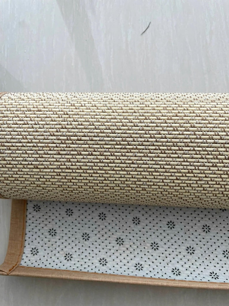 Pu&ograve; includere: Tappeto intrecciato beige e marrone arrotolato con un bordo marrone. Il lato inferiore presenta una superficie bianca con un motivo di piccoli punti neri e disegni floreali. Il tappeto &egrave; parzialmente srotolato, rivelando la sua trama e il suo design.