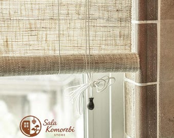 Puede incluir: Una persiana enrollable de bambú tejido en color beige con textura horizontal visible, que incluye un cordón blanco y un peso cilíndrico marrón oscuro. El logotipo de Sala Komorebi Store aparece abajo a la izquierda en un emblema circular.