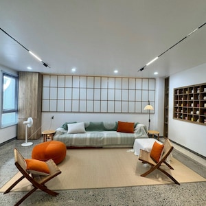 Pu&ograve; includere: Un moderno soggiorno con una palette di colori neutri. Include un divano a righe verdi e bianche con cuscini arancioni e bianchi, due sedie in legno con cuscini arancioni e un pouf arancione. Sono visibili anche un grande tappeto e un pannello a parete a griglia.