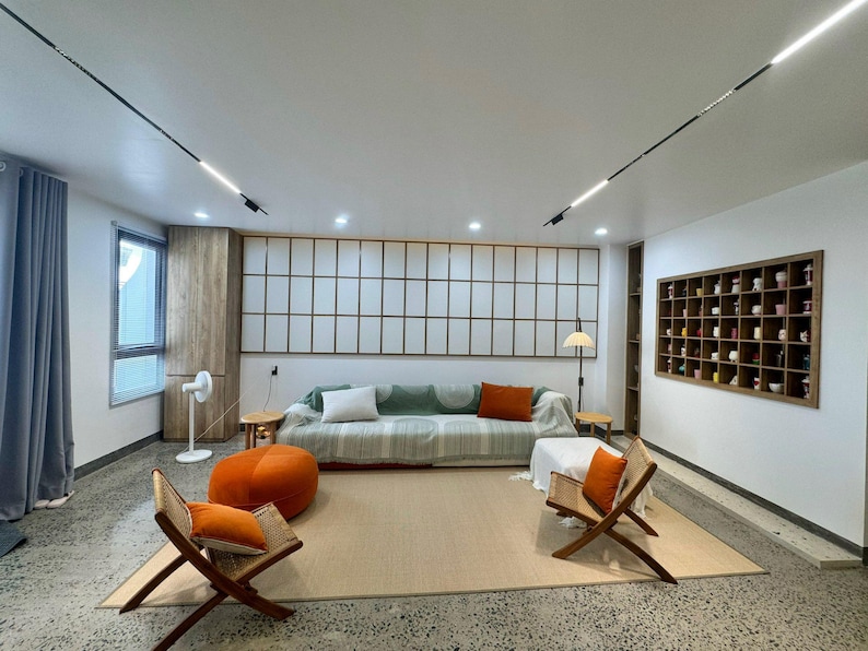 Pu&ograve; includere: Un moderno soggiorno con una palette di colori neutri. Include un divano a righe verdi e bianche con cuscini arancioni e bianchi, due sedie in legno con cuscini arancioni e un pouf arancione. Sono visibili anche un grande tappeto e un pannello a parete a griglia.
