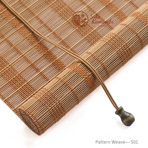 Può includere: Una tenda a rullo in bambù marrone chiaro con una trama intrecciata composta da sottili listelli verticali e fasce orizzontali più spesse. Un cordino sottile con un pomello metallico scuro a forma di campana pende dalla tenda. Il testo Sala Komorebi Store e Pattern Weave—S01 è visibile.
