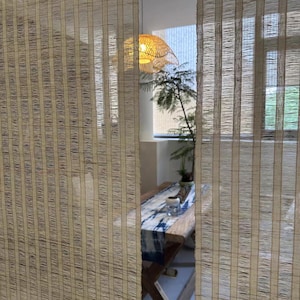 Puede incluir: Dos paneles verticales de cortinas de fibra tejida de color tostado claro, semitransparentes, con una sutil textura a rayas. Las cortinas cuelgan en primer plano, oscureciendo parcialmente la vista de una mesa de madera con un camino de mesa azul y blanco.