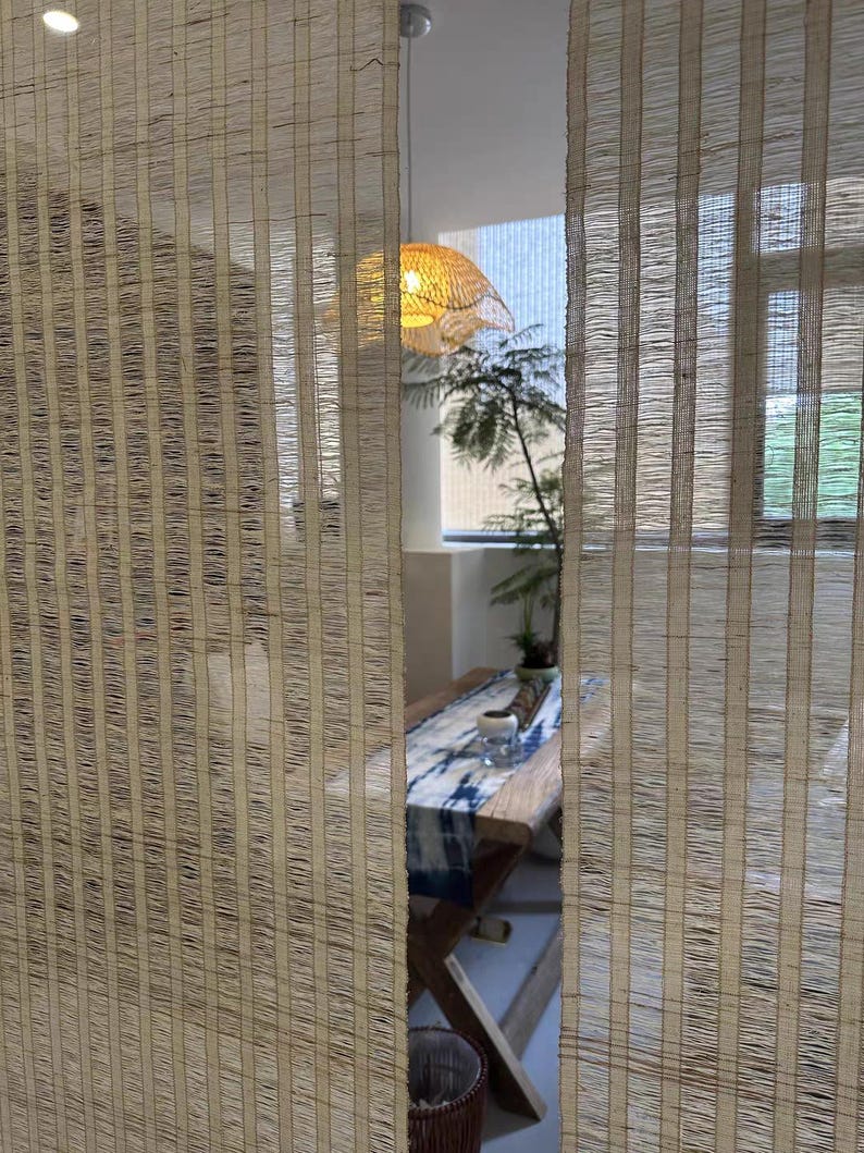 Puede incluir: Dos paneles verticales de cortinas de fibra tejida de color tostado claro, semitransparentes, con una sutil textura a rayas. Las cortinas cuelgan en primer plano, oscureciendo parcialmente la vista de una mesa de madera con un camino de mesa azul y blanco.