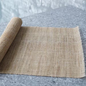Puede incluir: Un rollo de tela de arpillera de color tostado con una textura tejida visible y bordes deshilachados. El material es de color marr&oacute;n claro y presenta un patr&oacute;n de cuadr&iacute;cula consistente a lo largo de la tela, con un aspecto natural y fibroso.